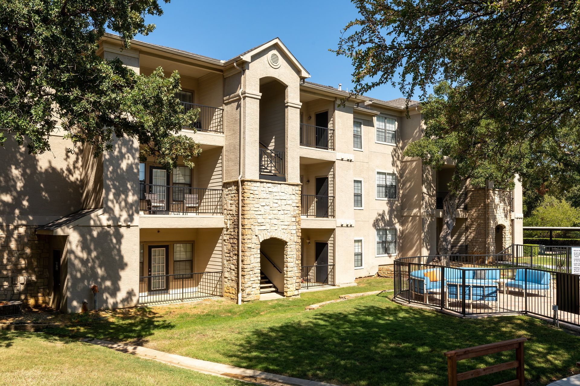 Apartment building exterior with a pool, surrounded by trees and green grass.