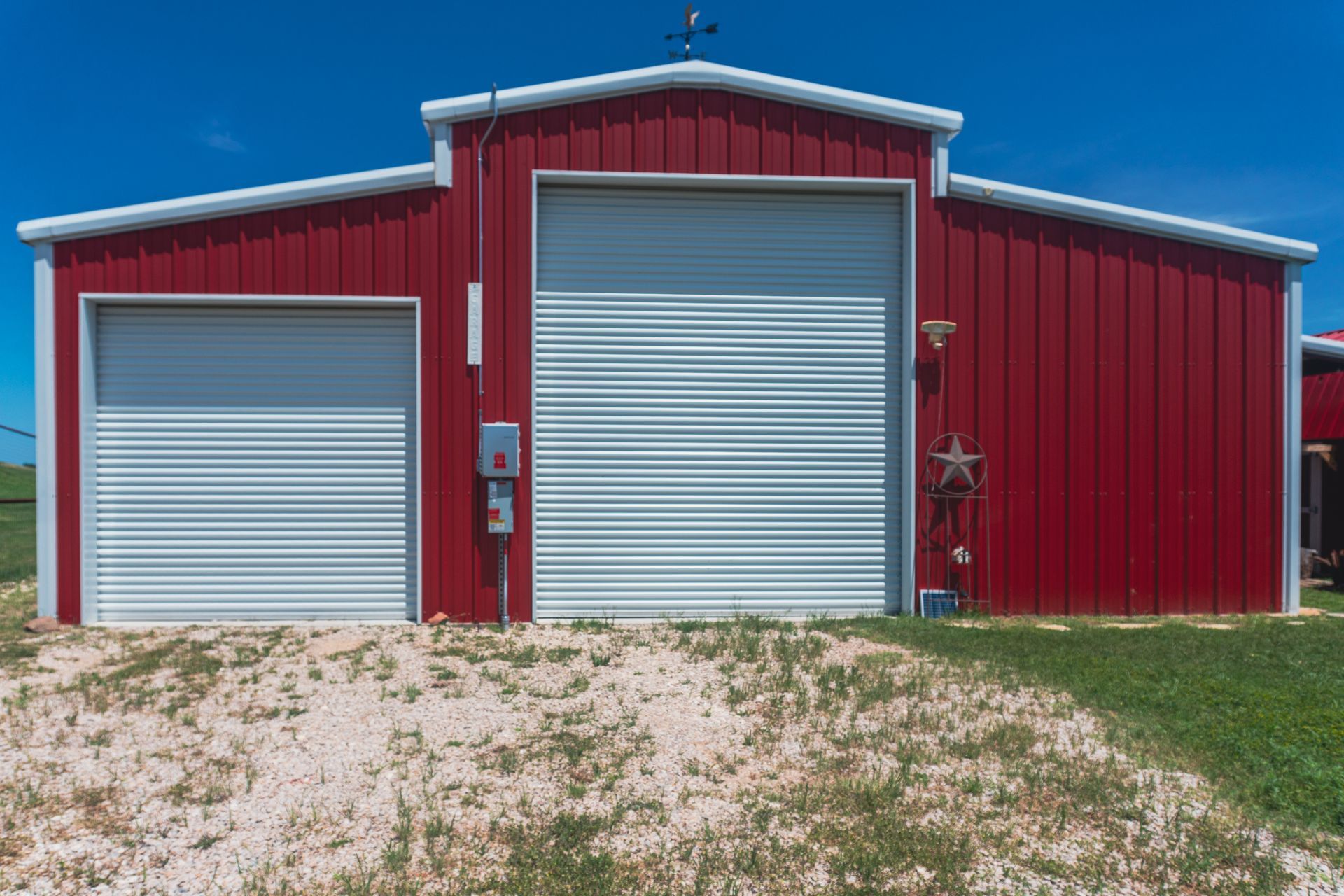 Red metal barn with two silver roll-up doors on a gravel and grass lot under a blue sky