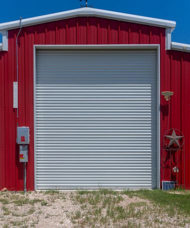 Red metal building with a large gray roll-up garage door and gravel foreground.