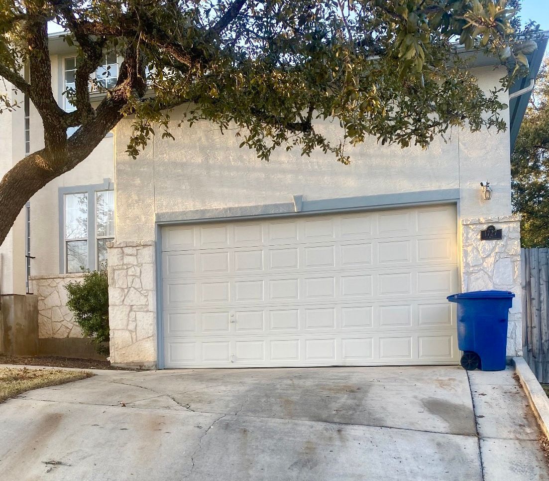 Suburban house with a white garage door, tree overhead, and a blue recycling bin by the driveway.