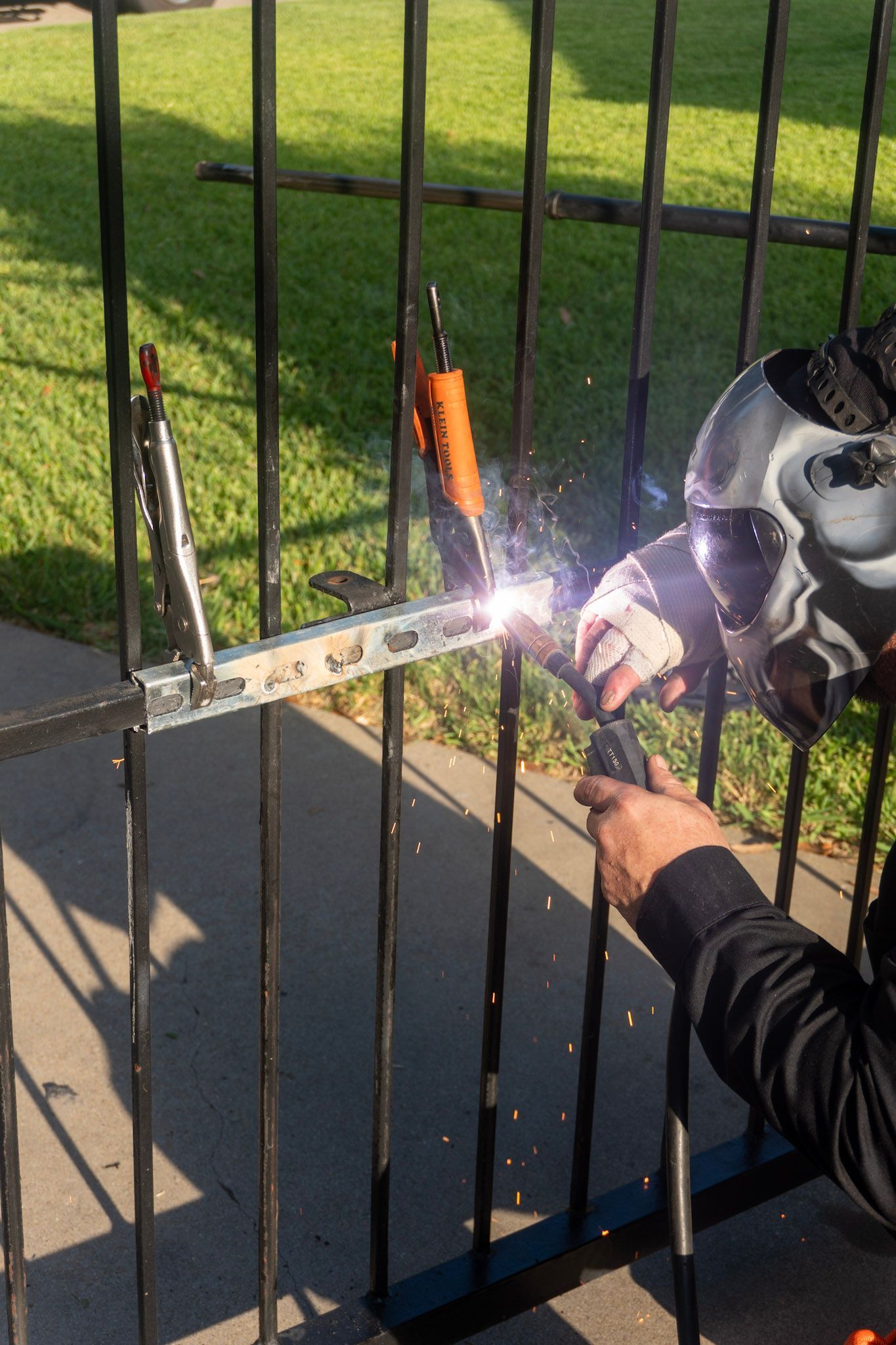 Person welding a black metal fence outdoors, with sparks flying near a grassy yard.
