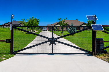 Black metal driveway gate with solar panels, set in front of a suburban house and lawn.