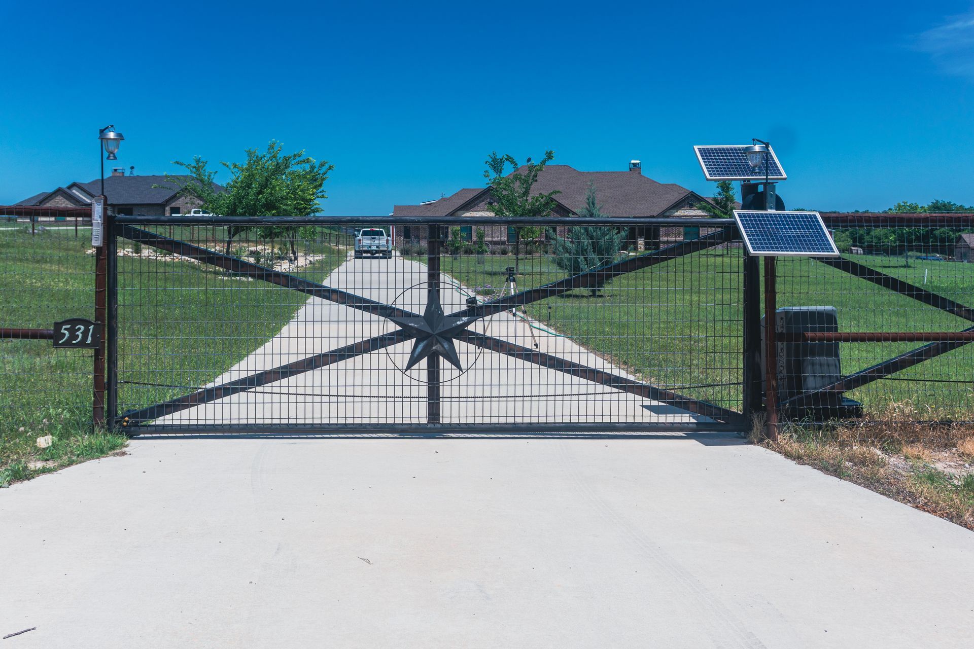 Black metal driveway gate with solar panels in front of a house under a blue sky.