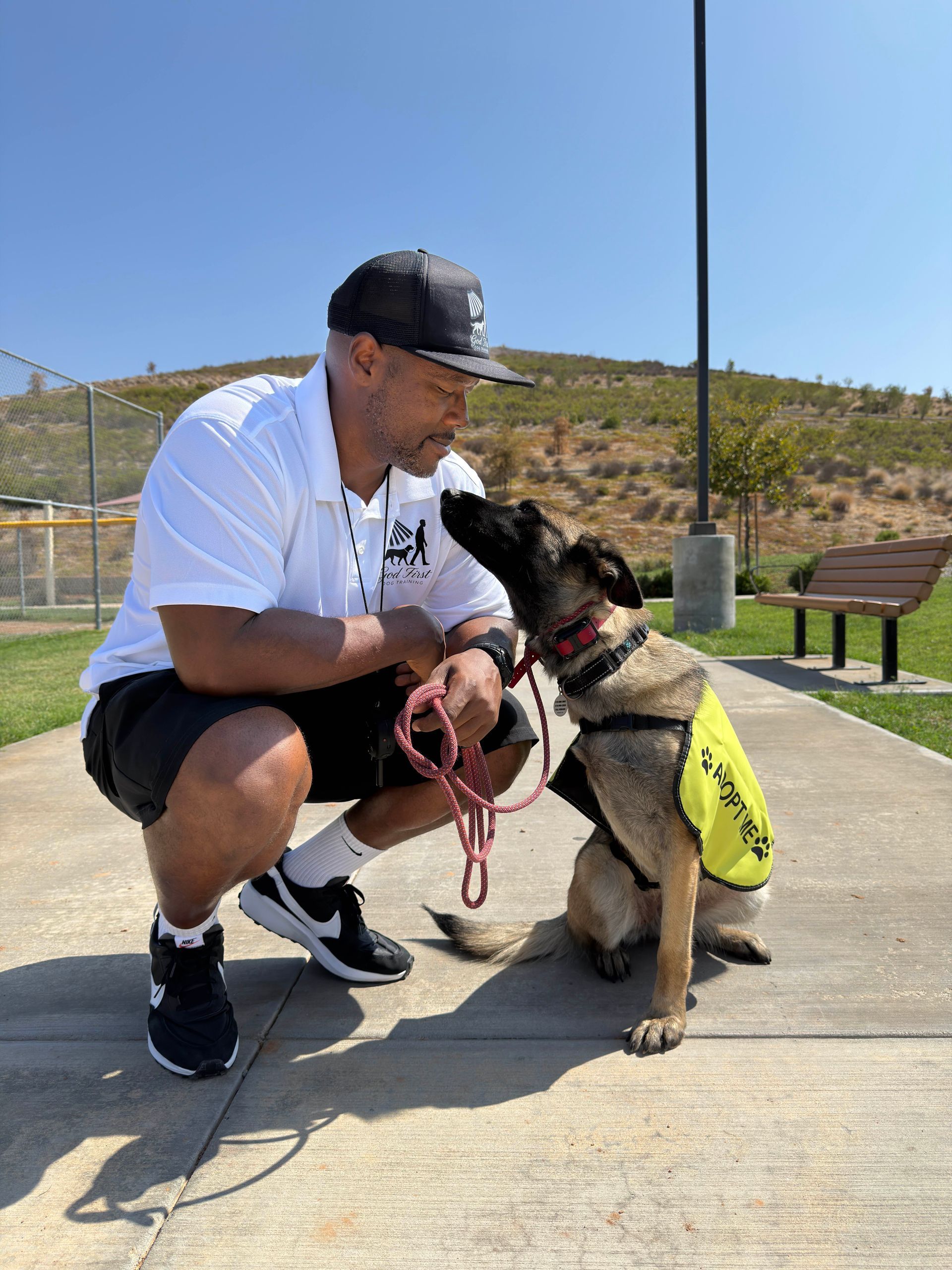 Man in cap and dog, both on a pathway. Dog wearing a vest looks up at the man, who is holding a leash. Sunny outdoor setting.
