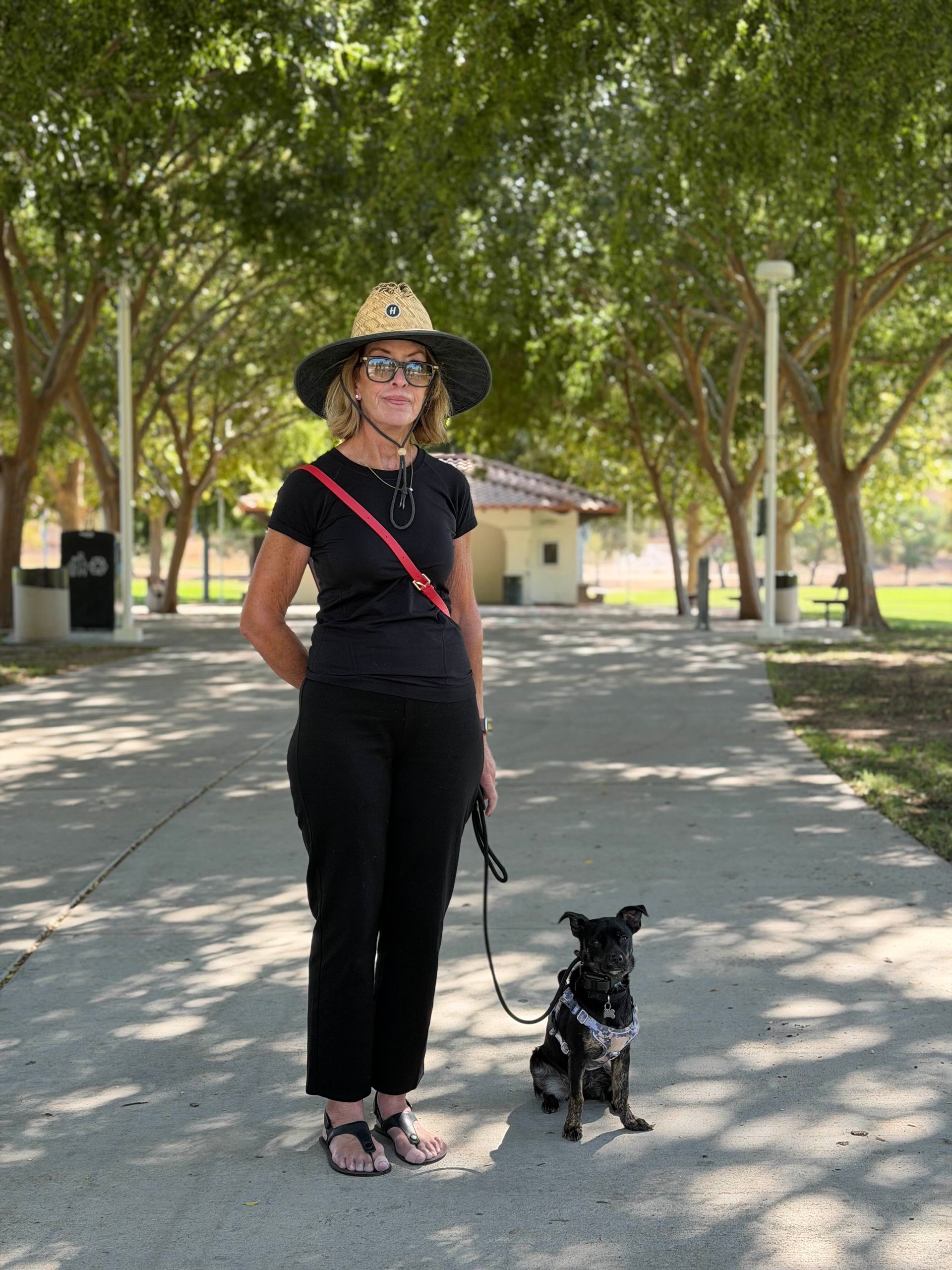 Woman in black outfit and hat with a dog on a leash in a park.