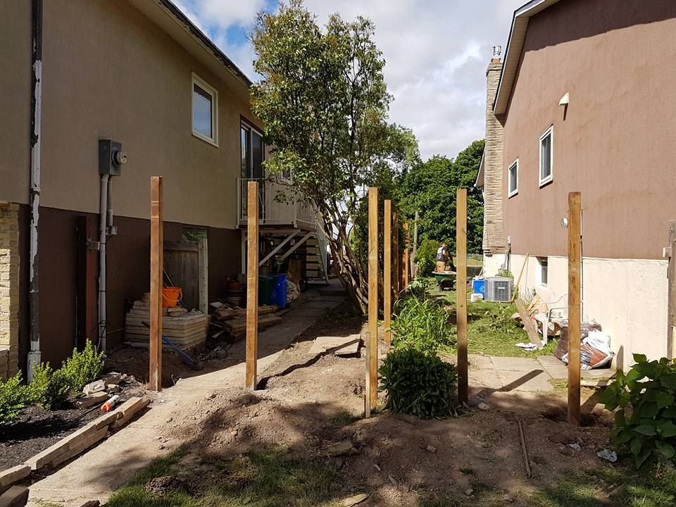 Wooden fence posts installed between two buildings, in a dirt and grass backyard under a cloudy sky.