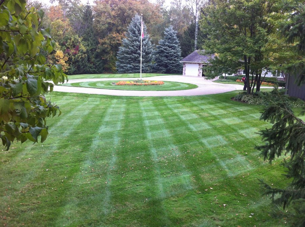 Lawn mowed in a checkered pattern, with a circular driveway and flowerbed around a flagpole, in a residential setting.