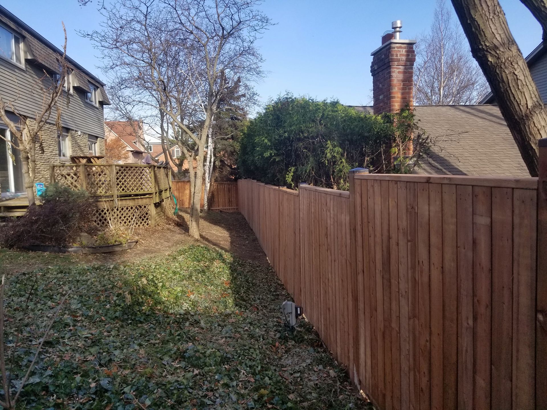 A brown wooden privacy fence borders a yard with a wooden deck and bare trees under a clear blue sky.