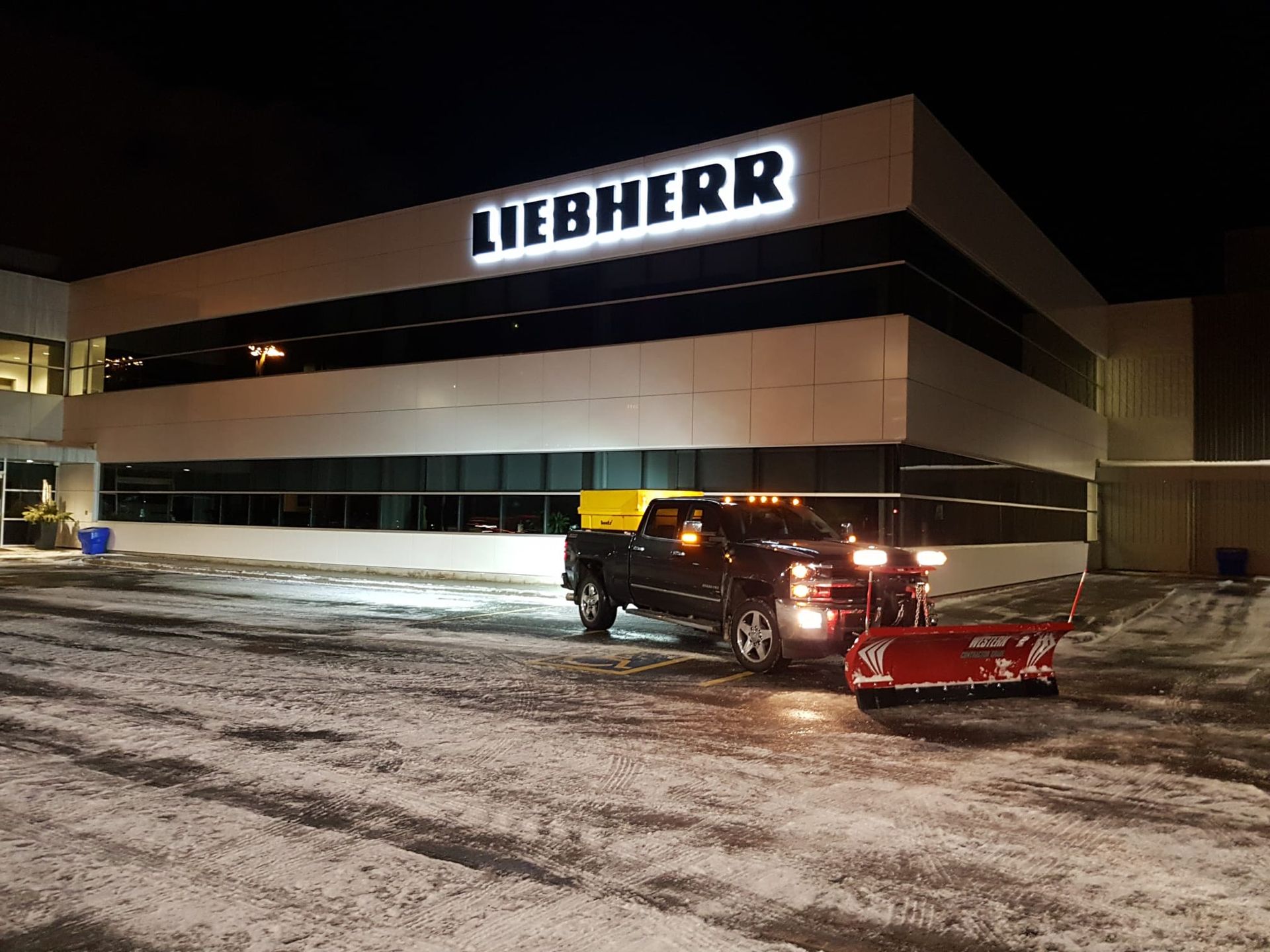 A truck is plowing snow in front of a liebherr building at night.