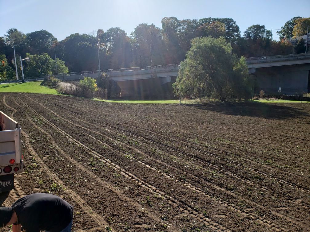 A person in the foreground tills a large, freshly plowed field beneath a highway bridge on a sunny day.