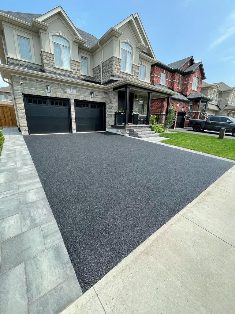 Two-story house with black asphalt driveway, stone trim and garage doors. Driveway bordered by grey pavers.