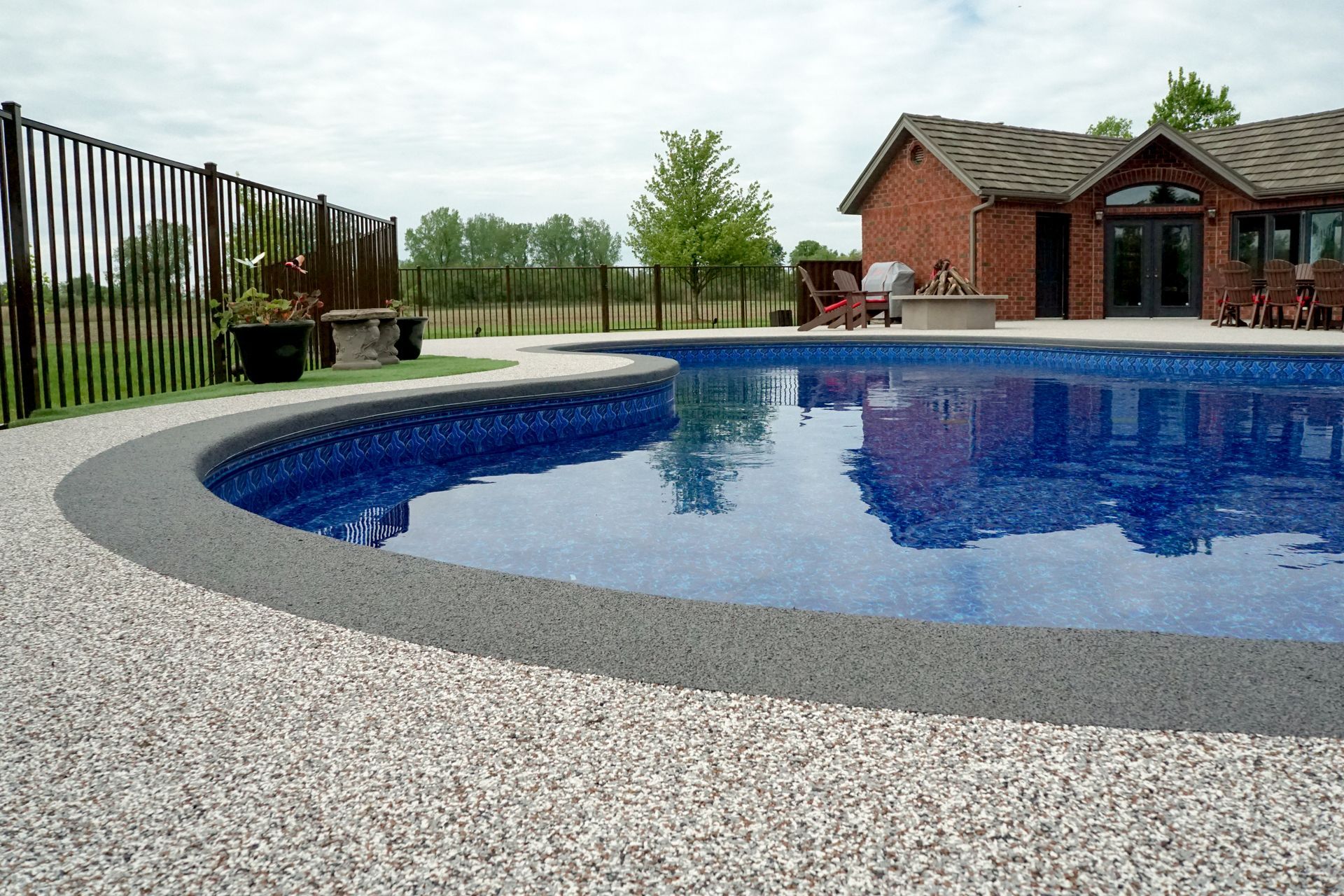 Pool with blue water, gray border, and light-colored pebbles. A brick house and black fence are in the background.