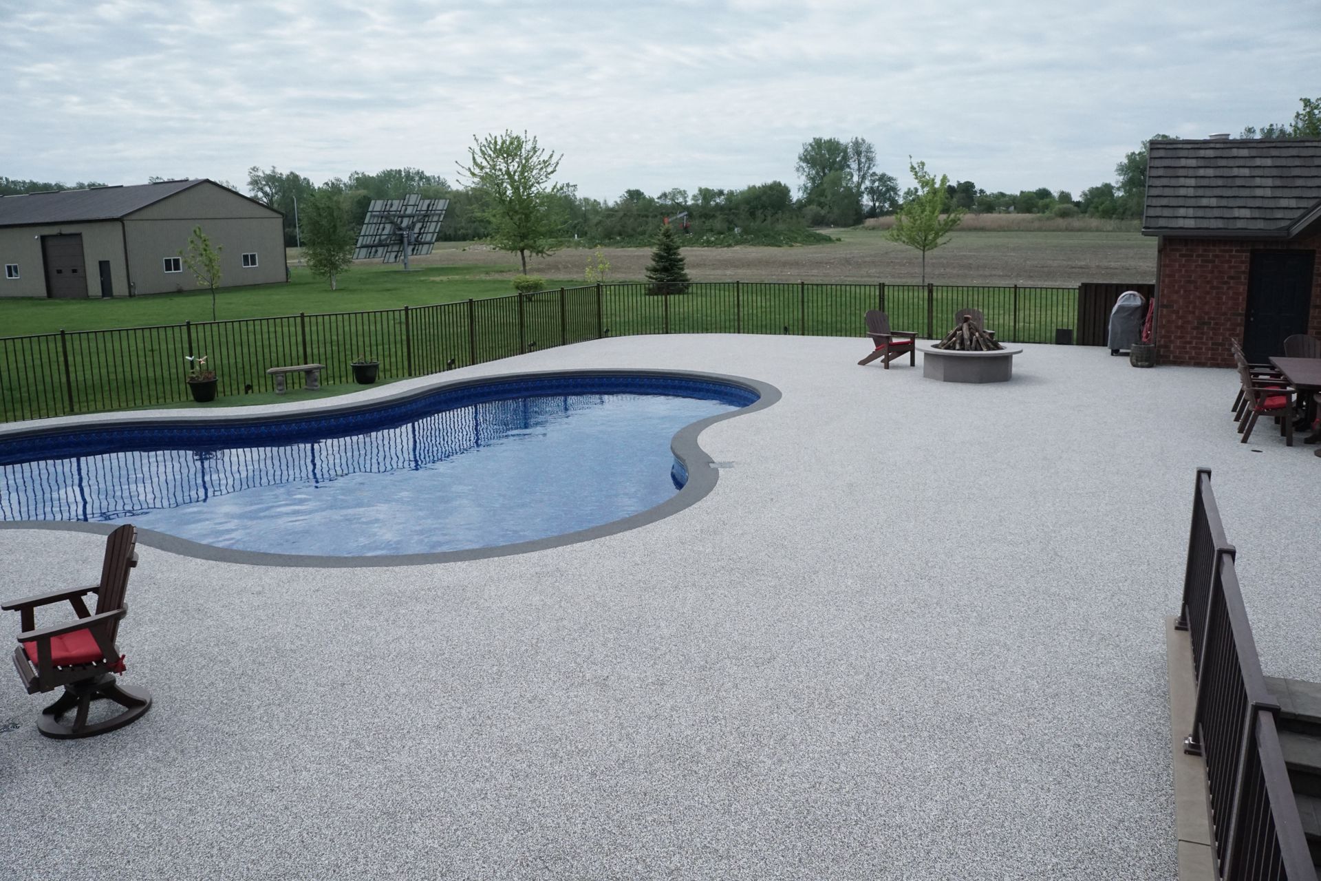 Pool and patio with a stone-like texture in a backyard setting. Cloudy sky and trees in background.