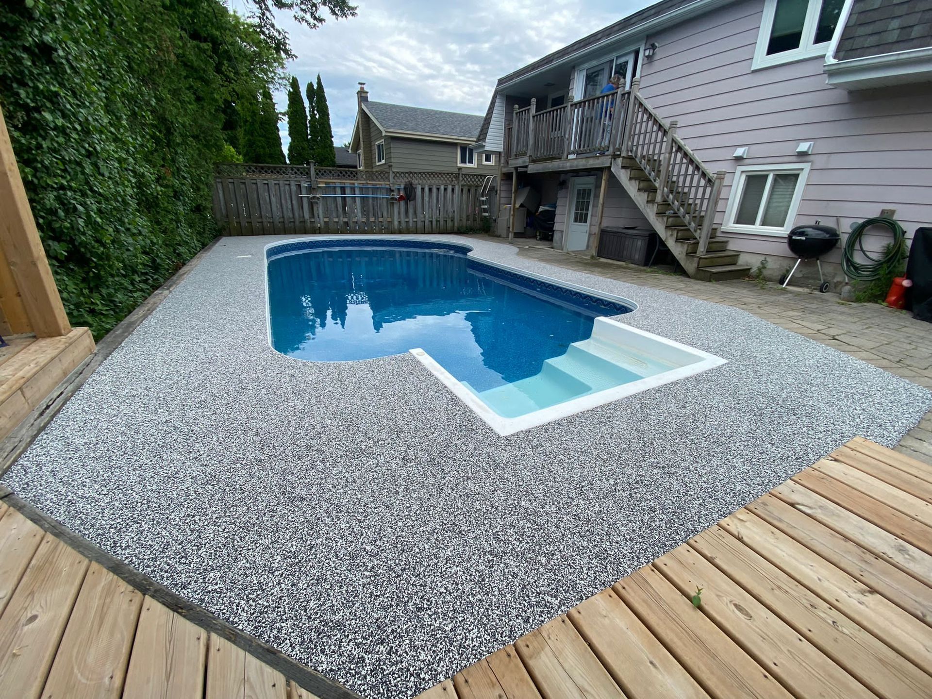 Backyard pool surrounded by pebble decking and wooden deck; gray siding house in background.