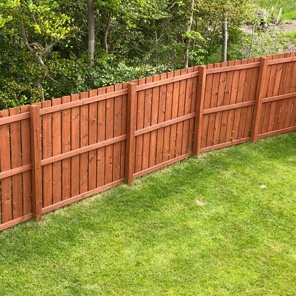 A wooden picket fence stained in a reddish-brown tone, situated in a grassy yard in front of green trees.