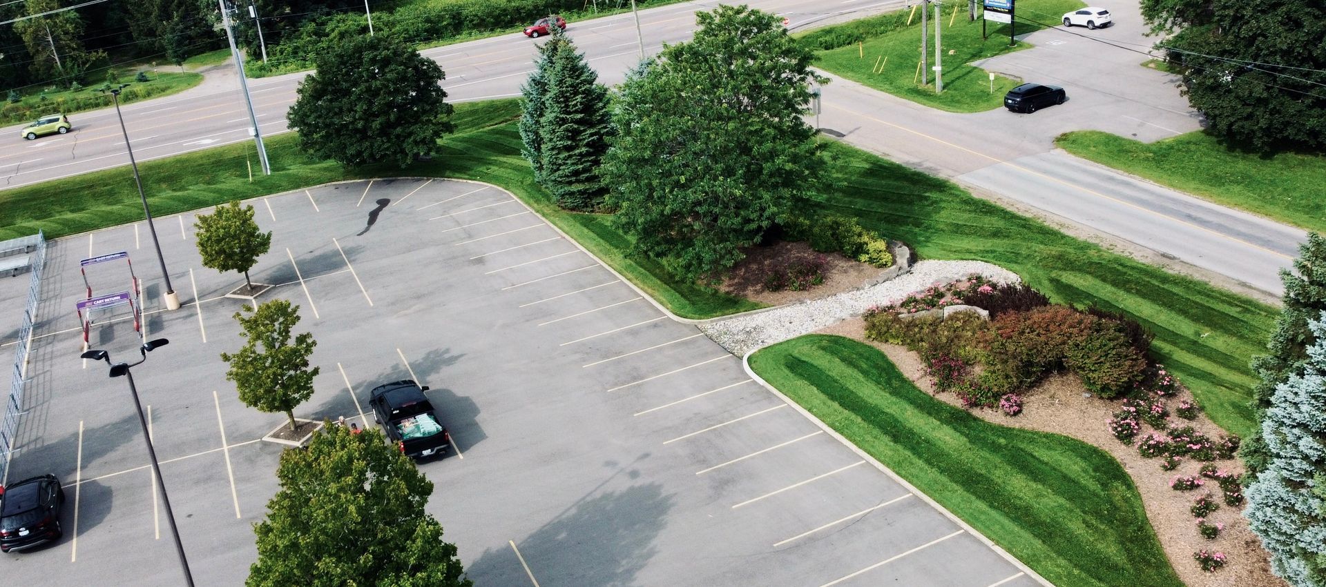 An aerial view of a parking lot, road, and green area. Black cars are parked.
