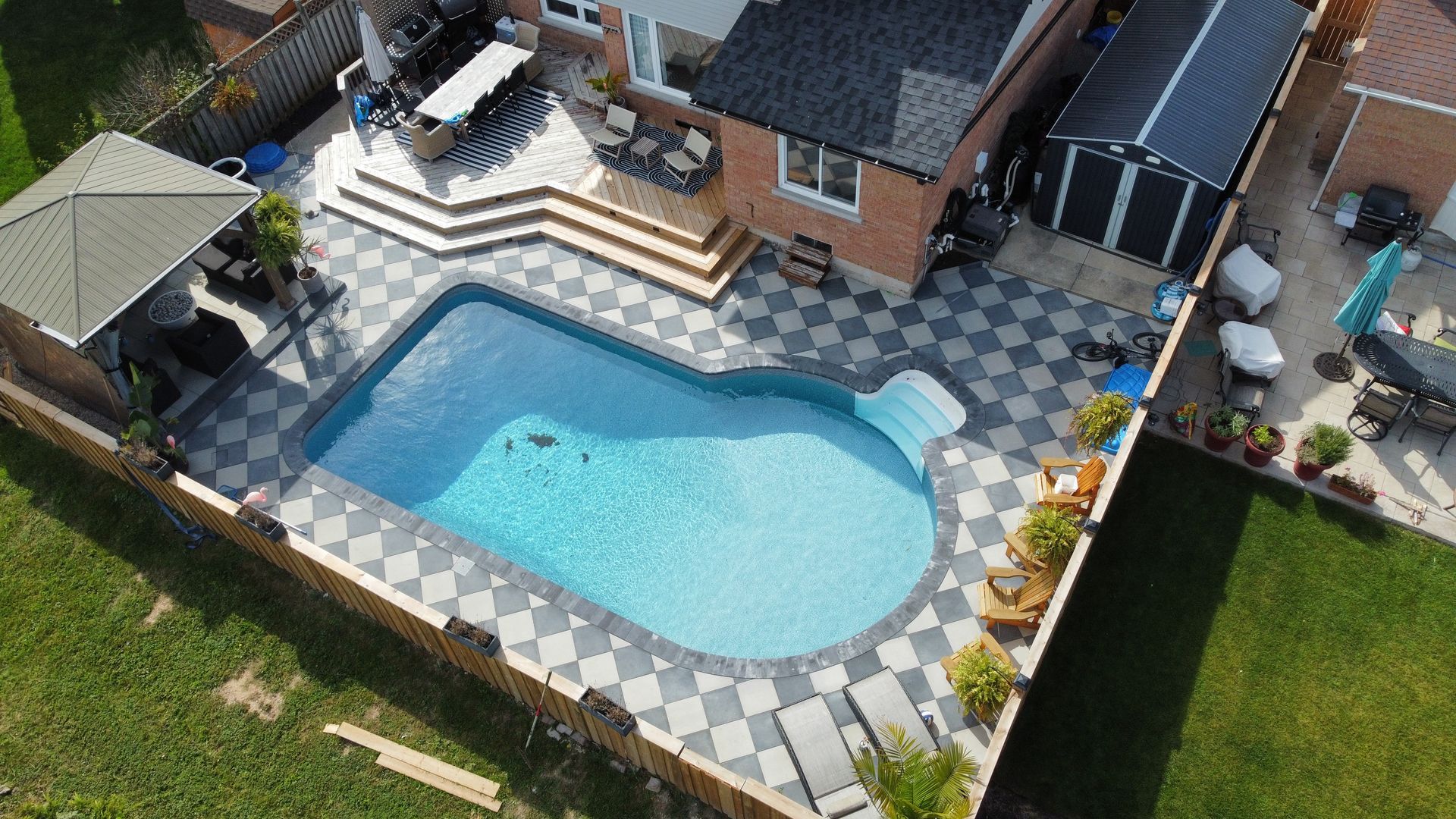An aerial view of a backyard featuring a swimming pool surrounded by a light and dark grey checkerboard patio and a gazebo.