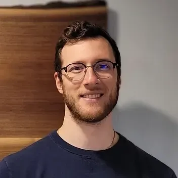 Man with glasses smiles, wearing blue shirt, against a wood-paneled wall.