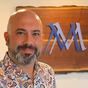 Bald man with beard smiles at the camera, stands in front of a wood sign with “RM” in blue and purple.