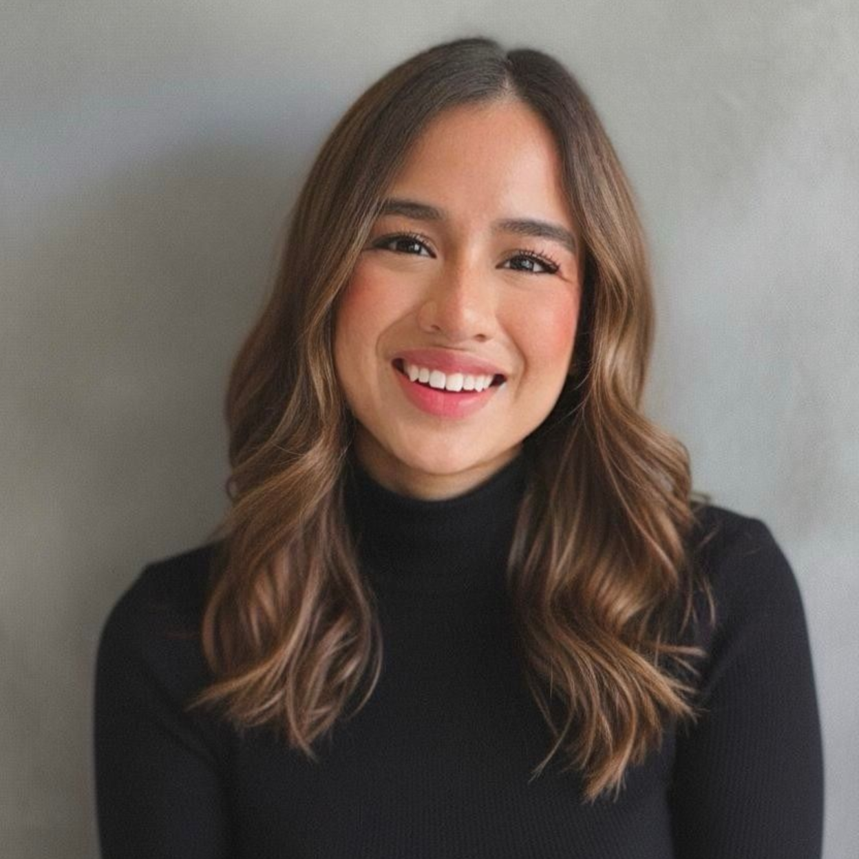 A person with long, wavy brown hair smiling while wearing a black turtleneck shirt against a gray background.