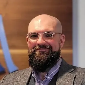 Bald man with glasses and full beard, smiling, wearing a blazer over a button-down shirt.