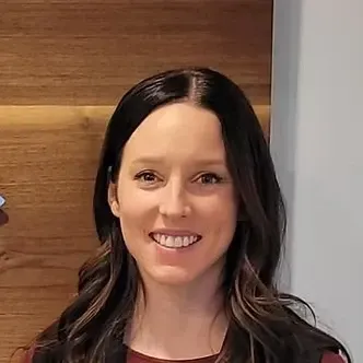 Woman with dark hair smiling, wearing a dark top, against a wood-paneled wall.