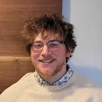 Man with glasses and beard smiles, wearing a cream sweater over a patterned shirt, set against a wood panel.