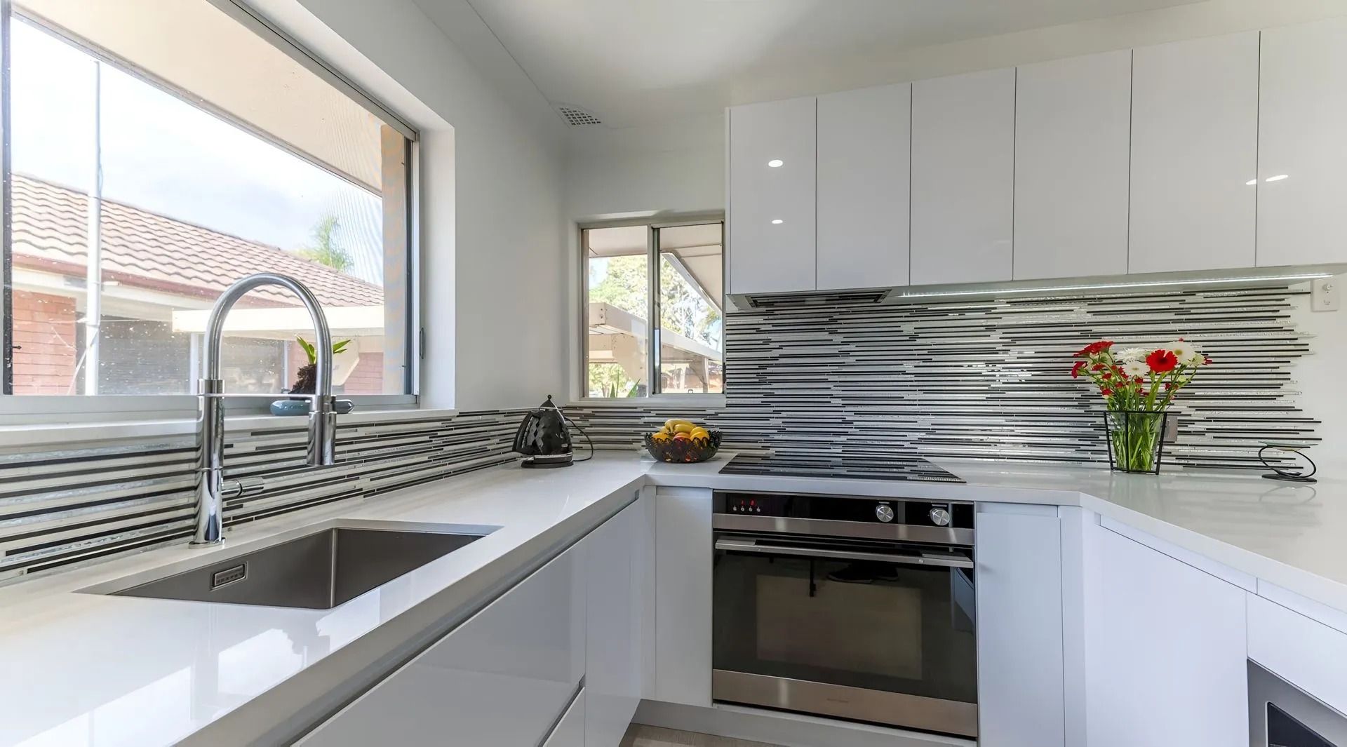 Modern white kitchen with a minimalist aesthetic, featuring a stainless steel sink, glossy cabinets, and textured tiles. — PKJ Design in Unanderra, NSW