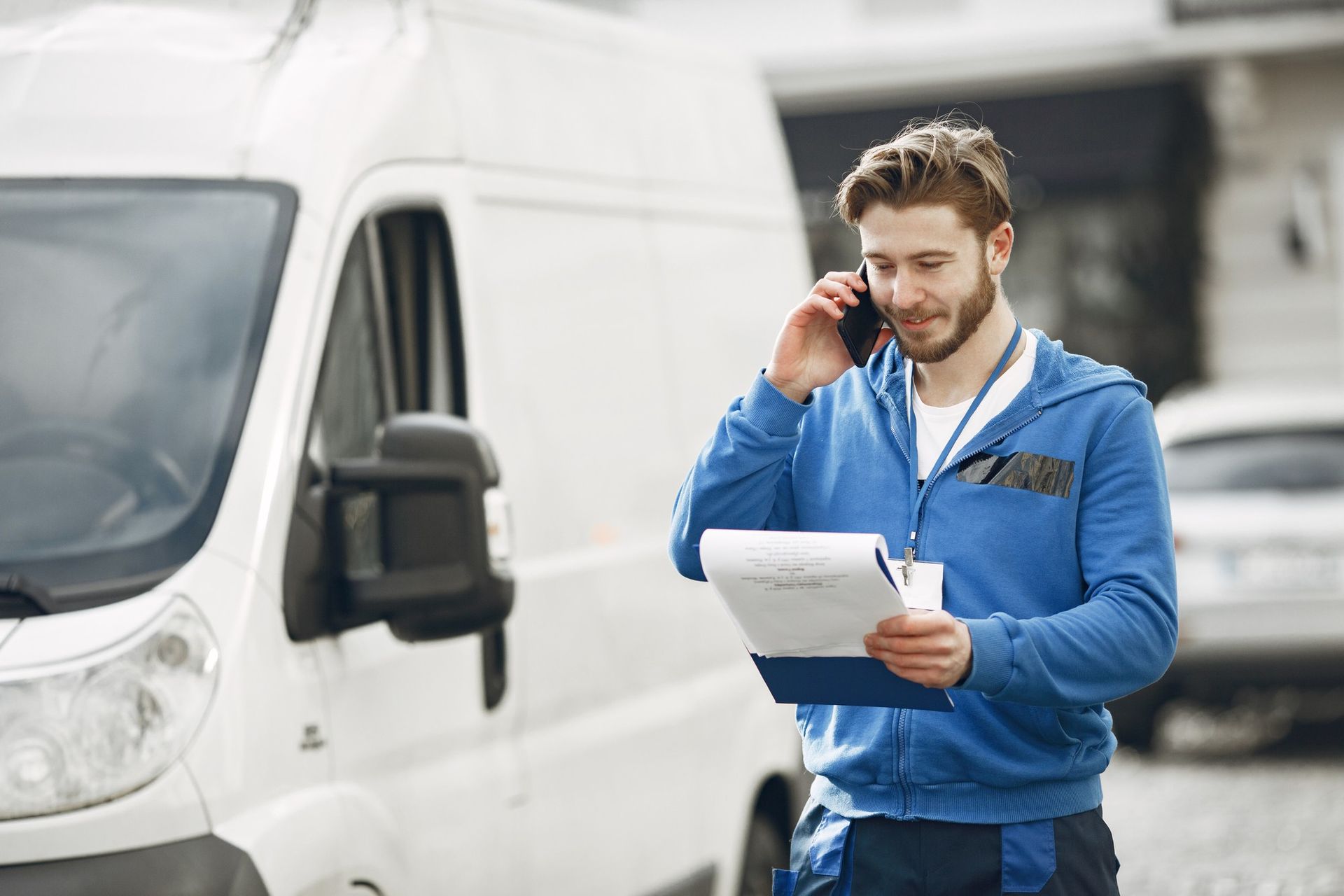 A man is talking on a cell phone while holding a clipboard in front of a van.