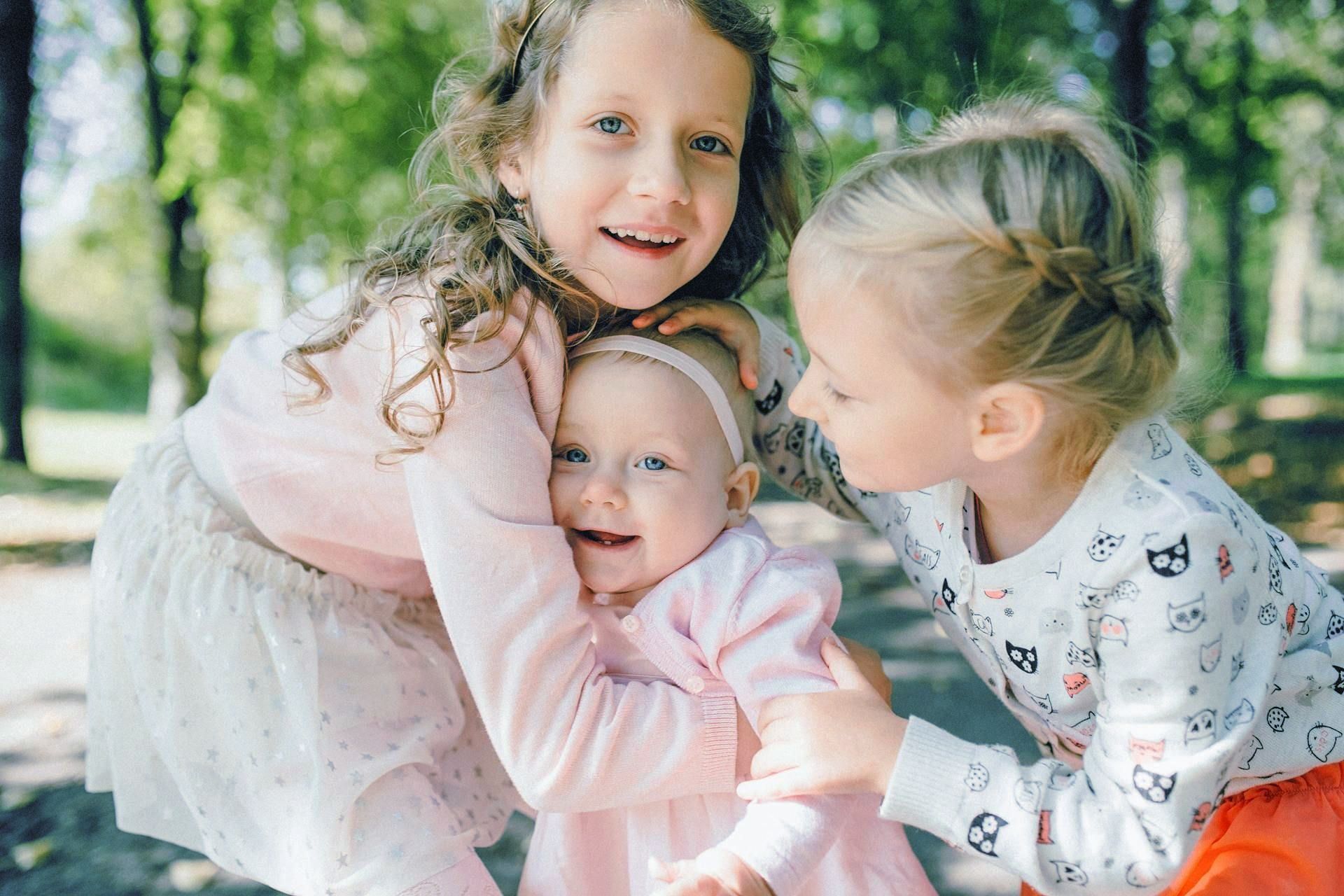 Two little girls are hugging a baby girl in a park.