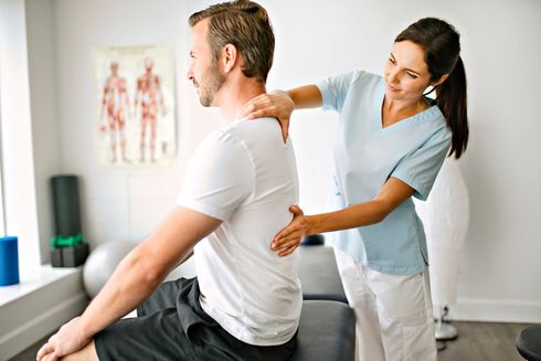 A woman is examining a man 's back while he sits on a table.