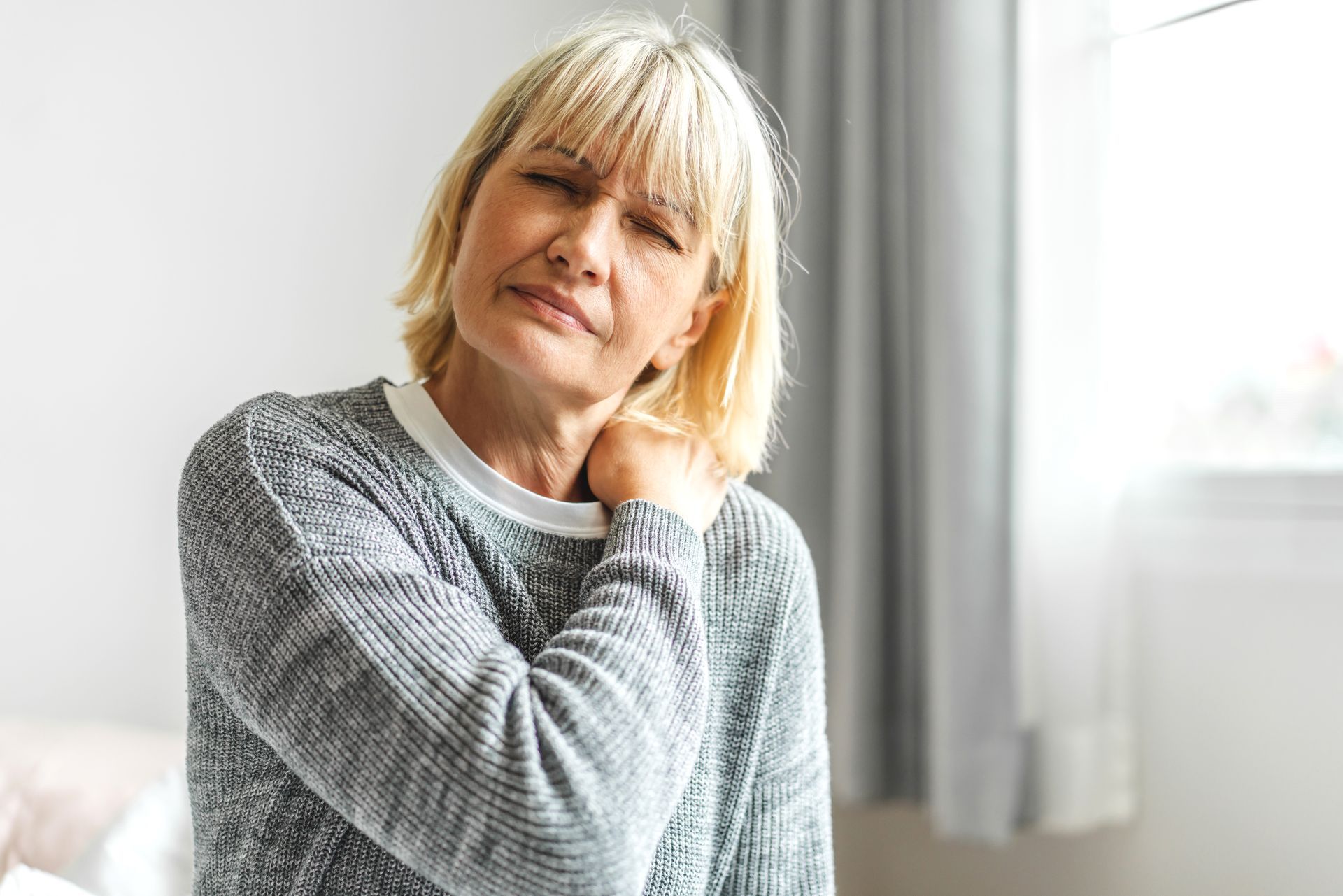 A woman is sitting on a bed holding her neck in pain.