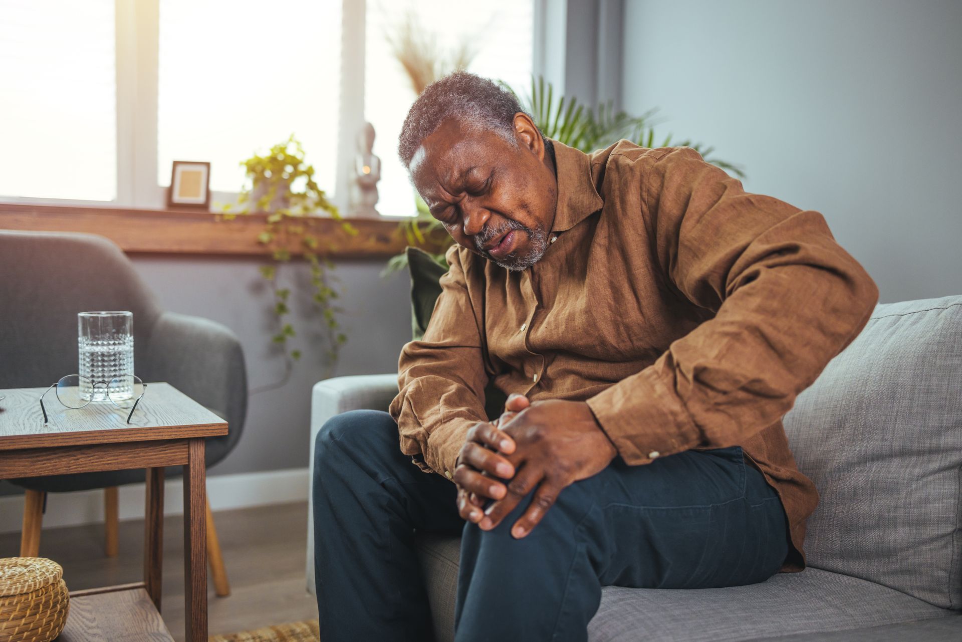 An elderly man is sitting on a couch holding his knee in pain.