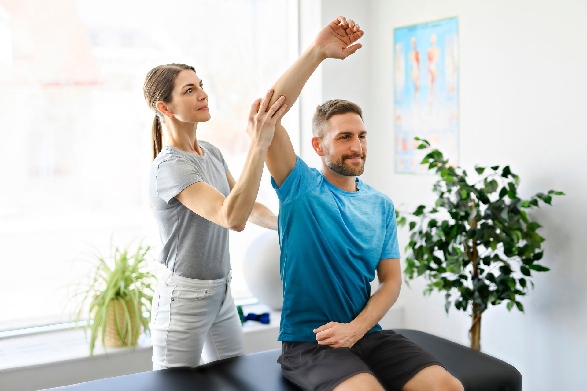 A man is sitting on a table while a woman stretches his arm.