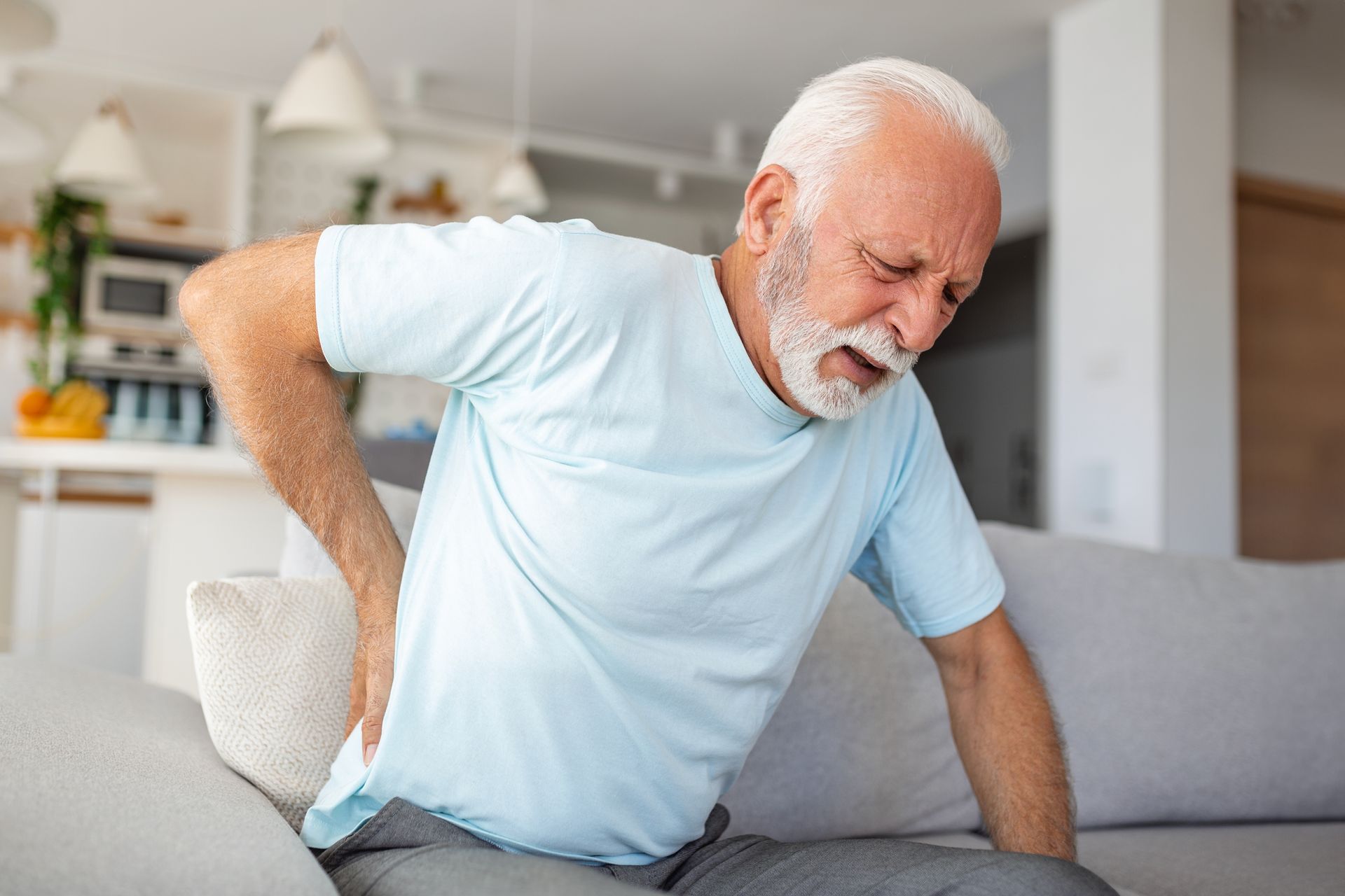 An elderly man is sitting on a couch holding his back in pain.