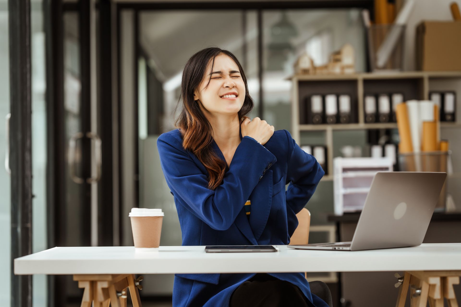 A woman is sitting at a desk with a laptop and holding her shoulder in pain.