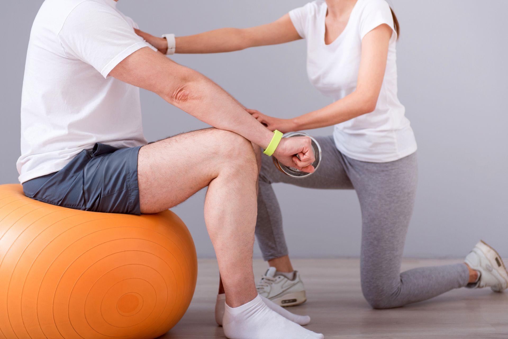 A woman is helping a man with his knee while sitting on an orange exercise ball.