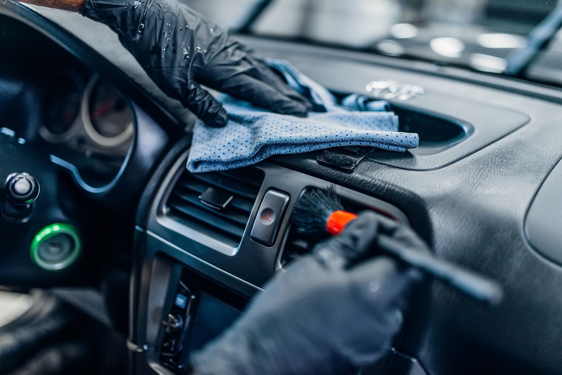Gloved hands use a soft brush and microfiber cloth to detail the air vents of a car dashboard.
