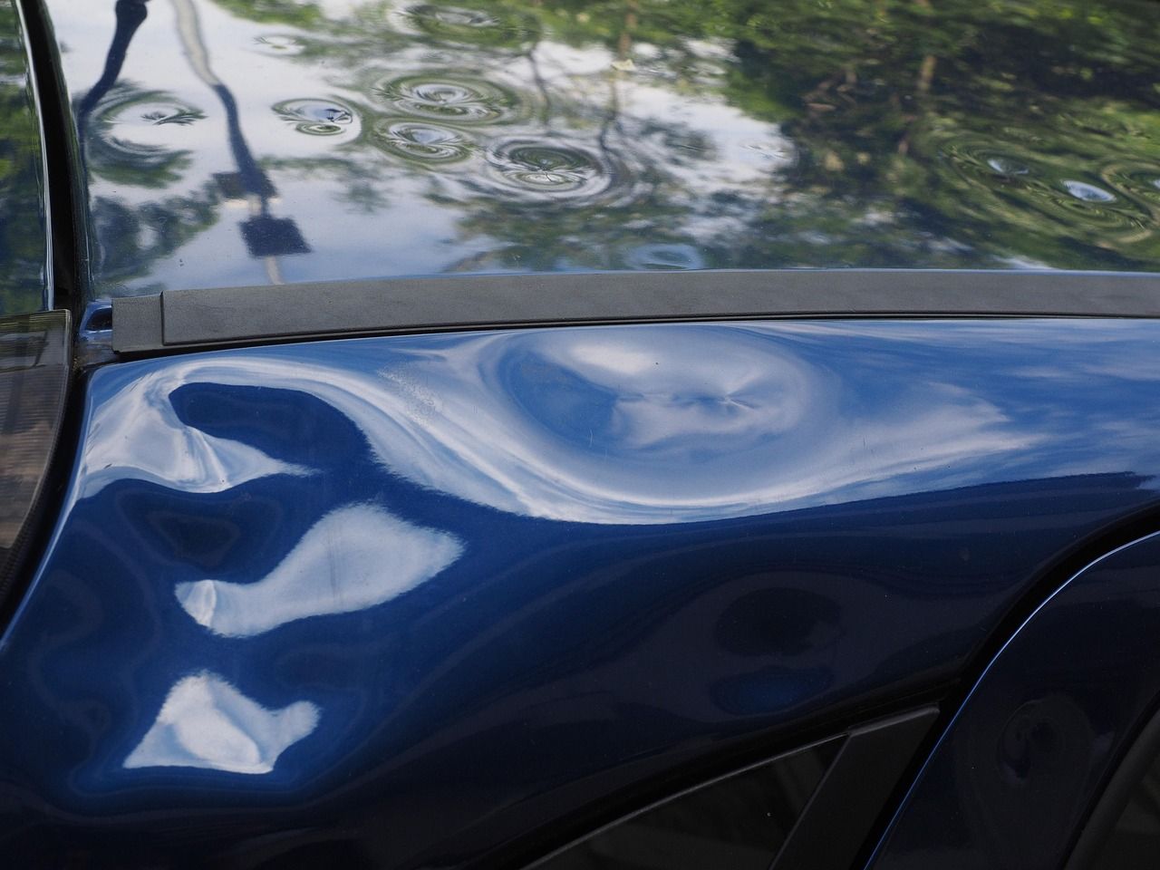 Multiple large dents on the side panel of a dark blue car, reflecting the sky and nearby trees.