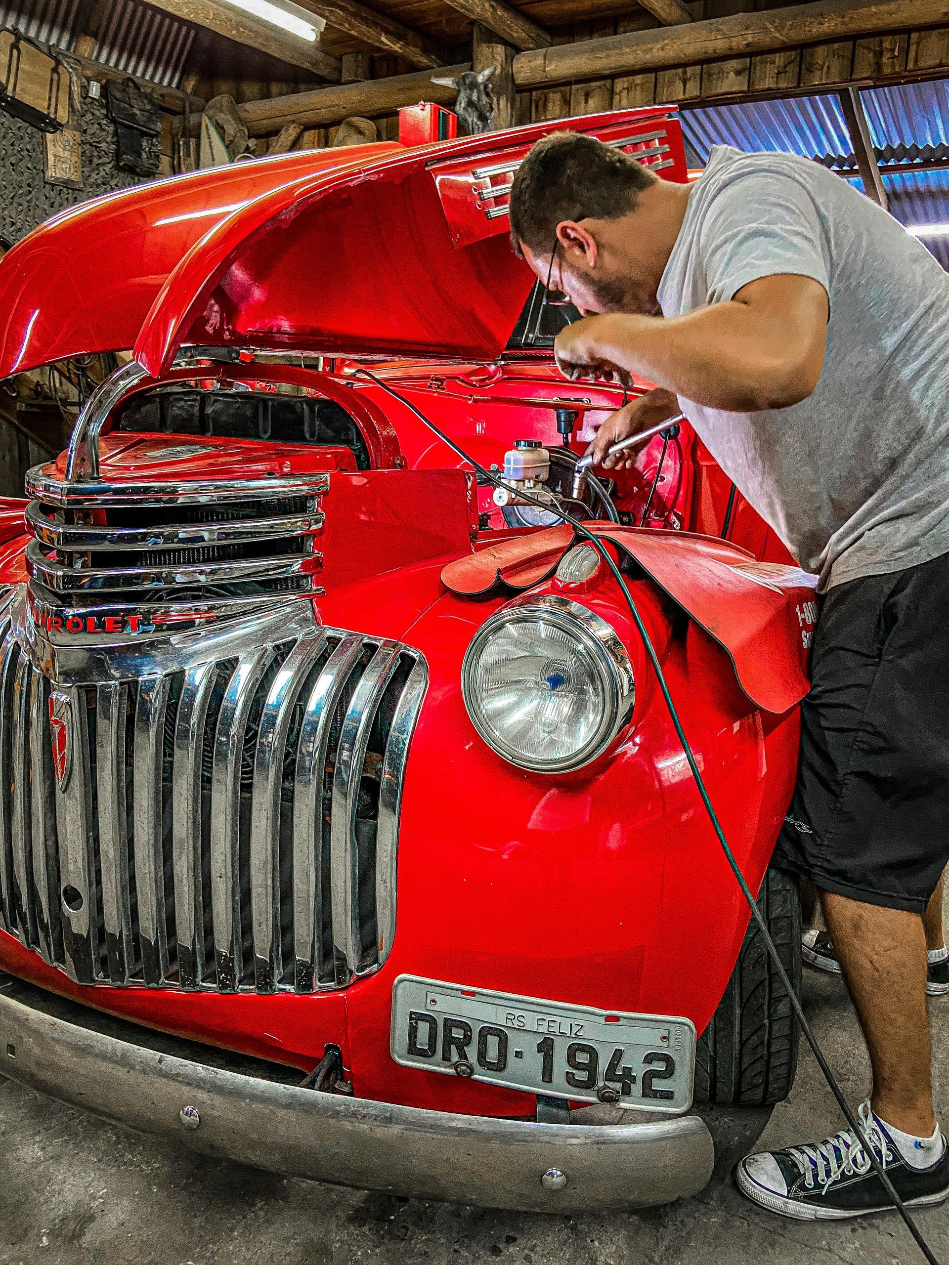 A person in a gray shirt works on the engine of a vintage red truck in a garage, holding a wrench under the open hood.