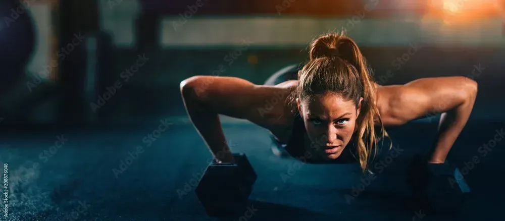 Woman doing push-ups on dumbbells in a gym. She has a determined expression. Blue and dark tones.