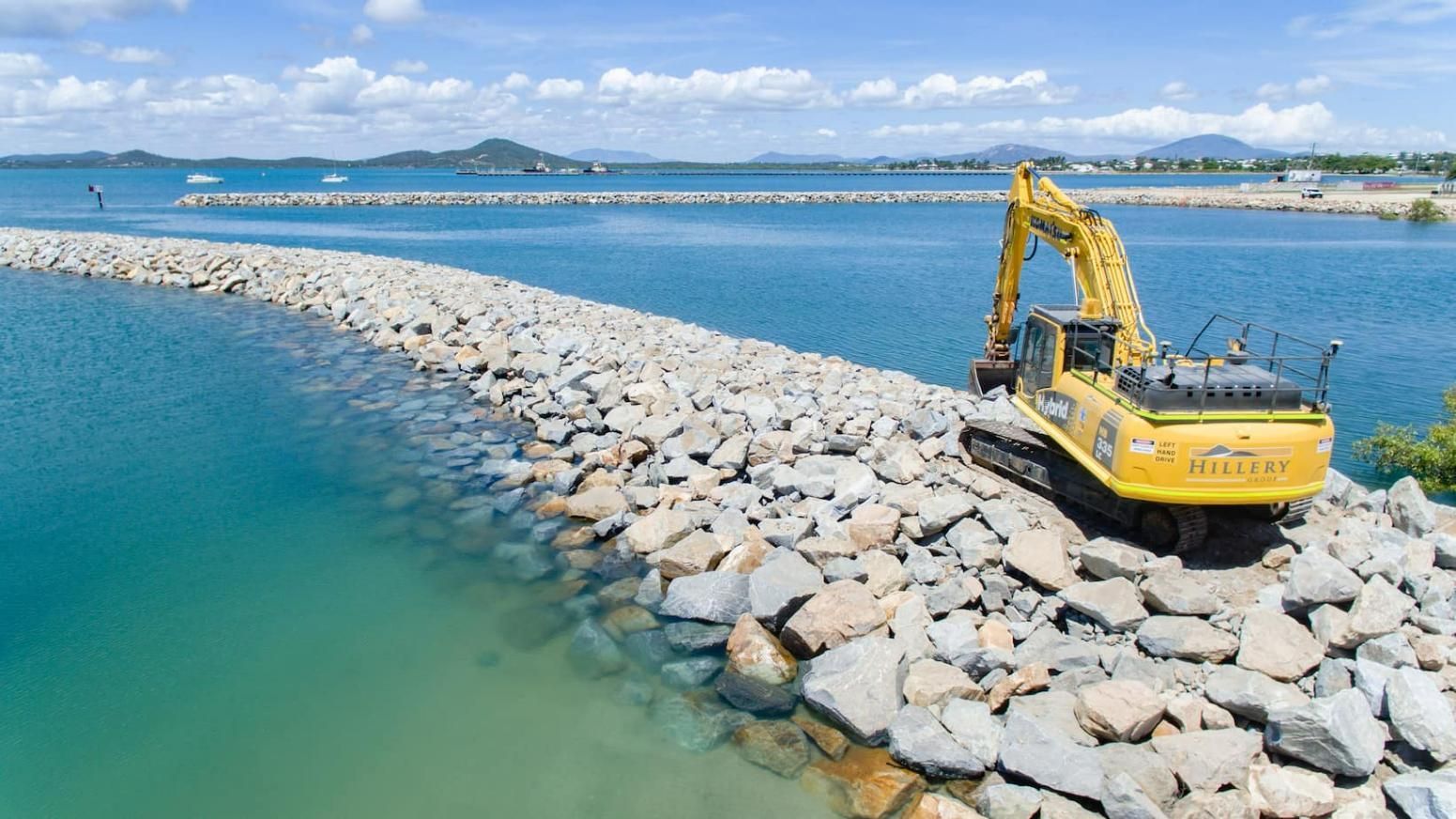 A Yellow Excavator Is Sitting on Top of A Pile of Rocks Next to A Body of Water — Hillery Group in Bowen, QLD