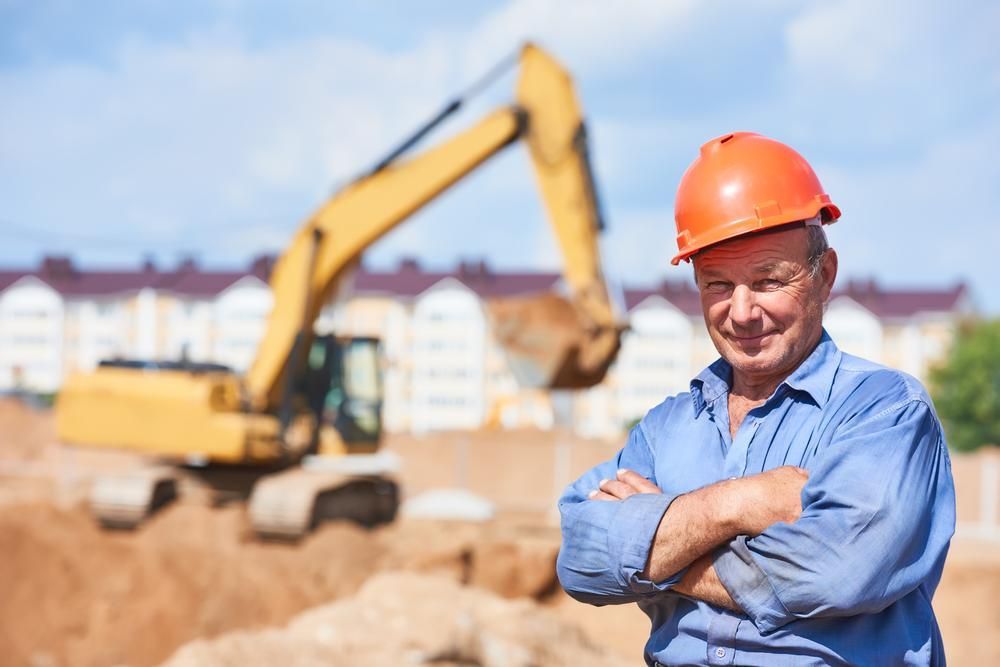 A Man Wearing a Hard Hat Is Standing on A Construction Site with His Arms Crossed — Hillery Group in Bowen, QLD