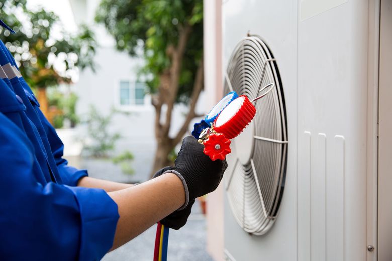 HVAC technician in blue overalls connecting gauges to an outdoor air conditioning unit.