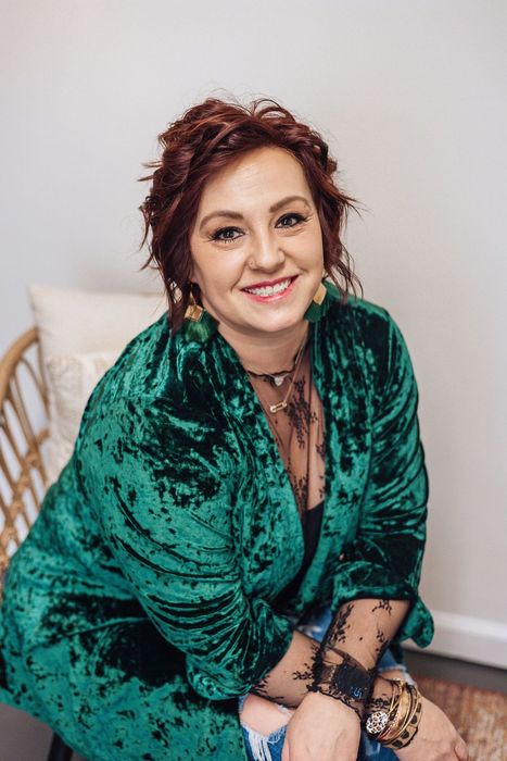 Woman in green velvet jacket smiles, seated in a wicker chair. Reddish-brown hair, visible jewelry.