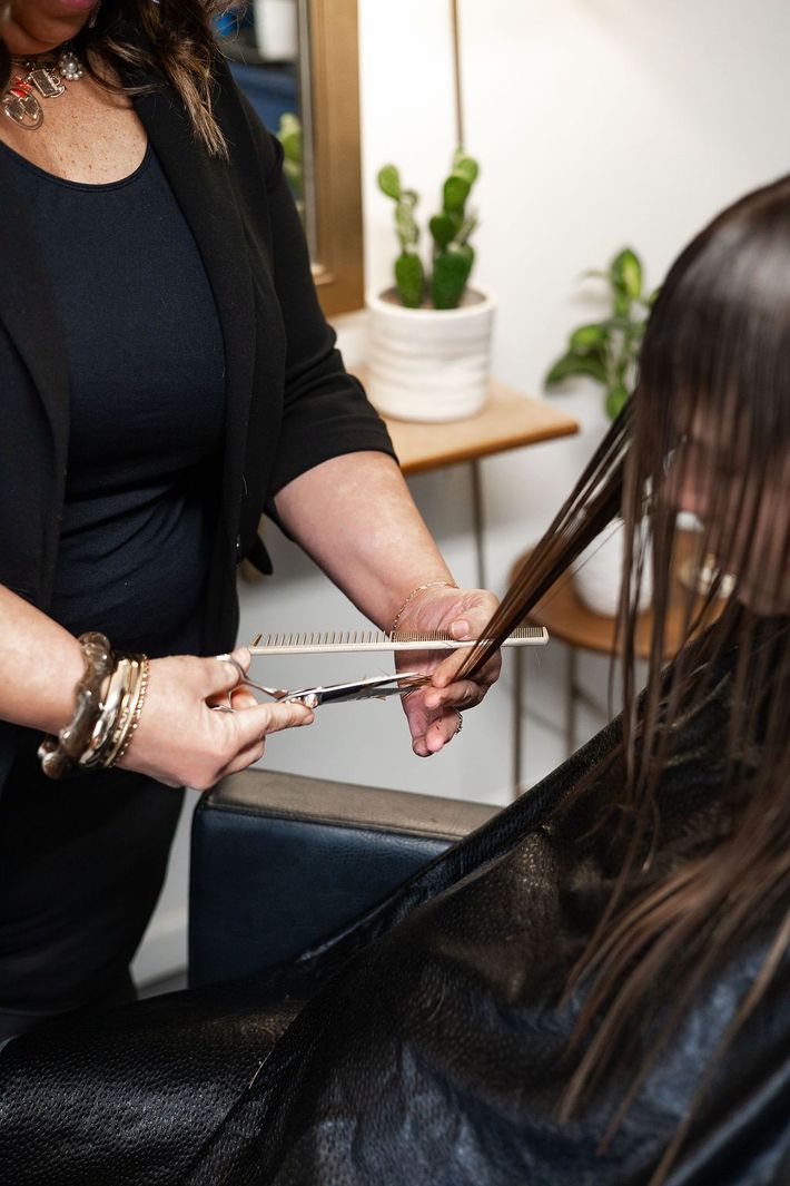 Hairdresser cutting client's wet, dark hair with scissors and a comb in a salon setting.