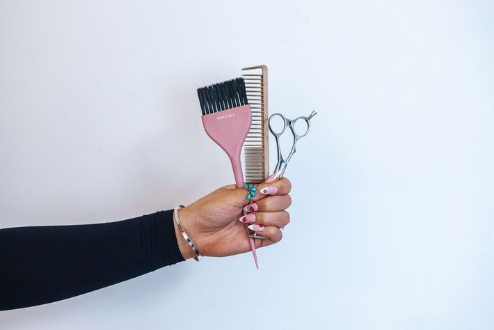 Hand holding pink hair dye brush, comb, and shears against white wall.