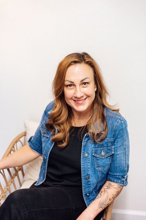 Woman smiling, sitting in wicker chair, wearing denim jacket and black top, against a white wall.