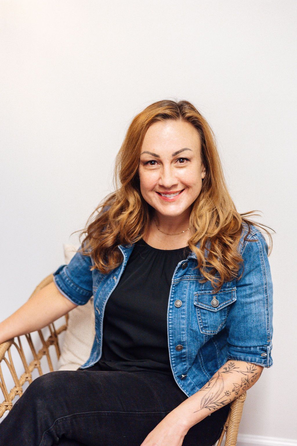 Woman seated in a wicker chair, smiling. Wearing a denim jacket, black top, and has an arm tattoo.