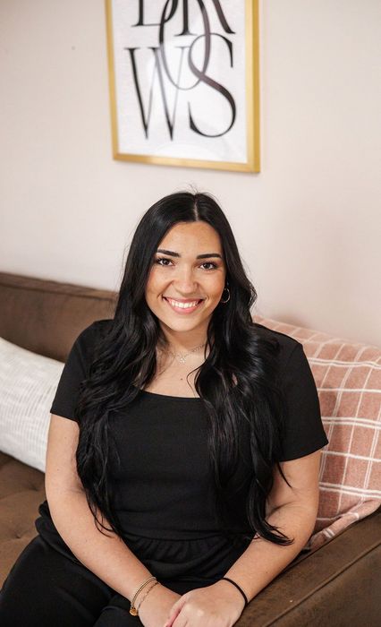 Woman with long dark hair smiles on a brown couch. A gold-framed print hangs on a light pink wall.